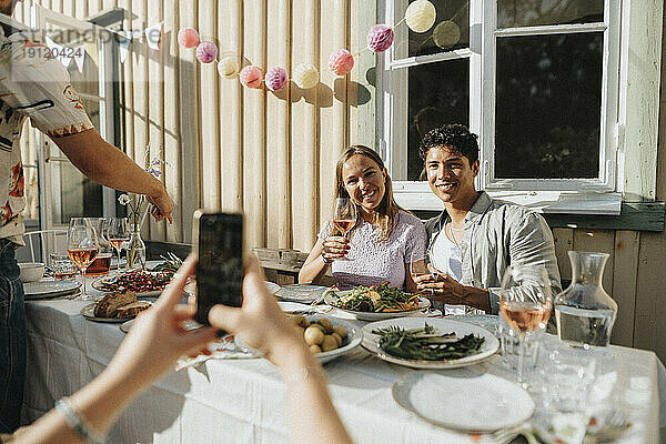 Frau  die ein Bild von männlichen und weiblichen Freunden anklickt  die ein Weinglas während einer Dinnerparty im Café halten