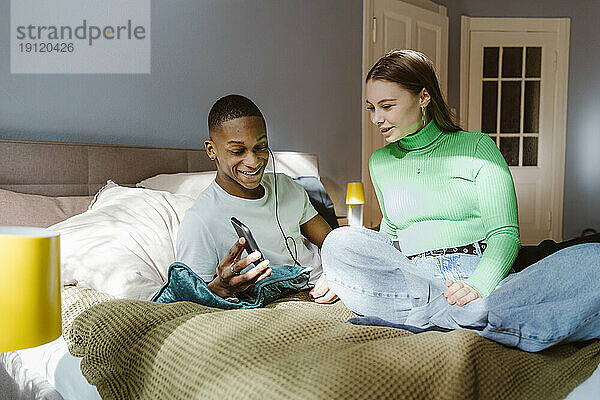 Lächelnder junger Mann teilt sein Smartphone mit einer Freundin auf dem Bett zu Hause