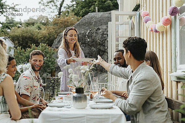 Lächelnde Frau  die einem männlichen Freund  der am Tisch sitzt  den Teller reicht  während einer Dinnerparty außerhalb eines Cafés