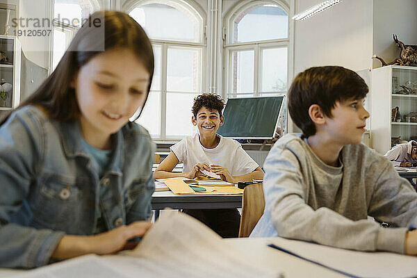 Porträt eines glücklichen Schuljungen  der mit Freunden im Klassenzimmer am Schreibtisch sitzt