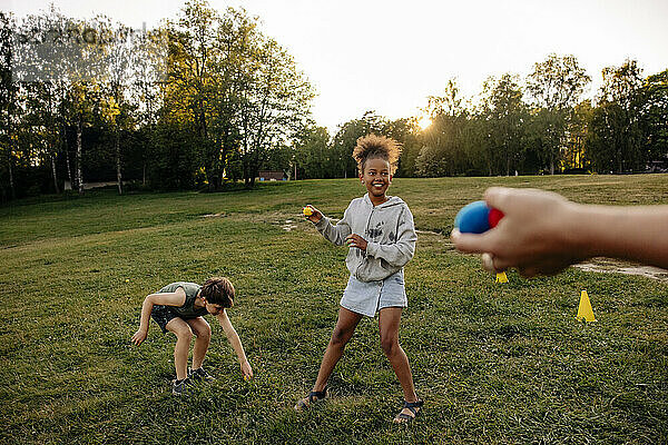 Lächelndes Mädchen  das mit Bällen spielt  während es neben einem männlichen Freund auf einem Spielplatz im Gras steht