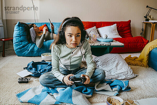 Frau spielt ein Videospiel  während ihre Freundin auf dem Sofa zu Hause ihr Smartphone benutzt