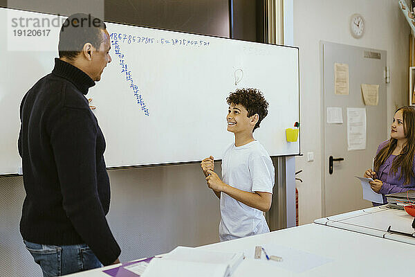 Lächelnder Junge im Gespräch mit einem männlichen Lehrer  der in der Nähe der Tafel im Klassenzimmer steht