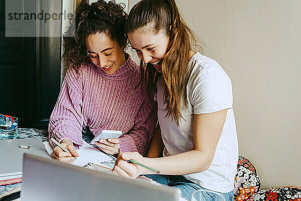 Glückliche junge Freundinnen benutzen ihr Smartphone bei den Hausaufgaben