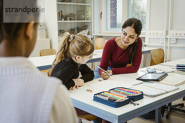 Lächelnde Lehrerin erklärt Schülerin am Schreibtisch sitzend im Klassenzimmer