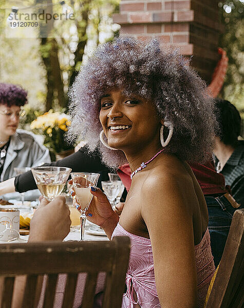 Seitenansicht Porträt der lächelnden Transfrau mit lockigem Haar sitzt mit Freunden während der Party im Hinterhof
