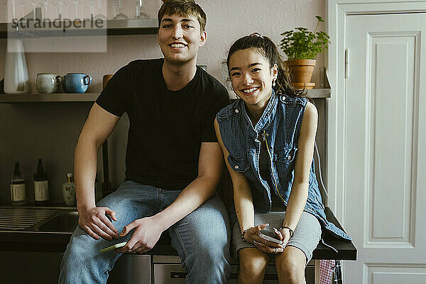 Porträt von lächelnden jungen Freunden  die zusammen auf dem Küchentisch zu Hause sitzen
