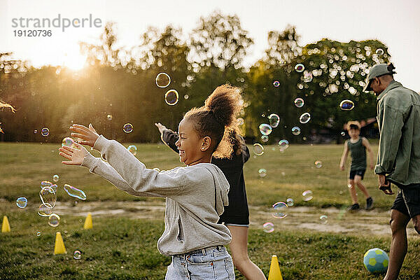 Seitenansicht eines glücklichen Mädchens  das Seifenblasen fängt  während es mit Freunden auf dem Spielplatz im Ferienlager spielt