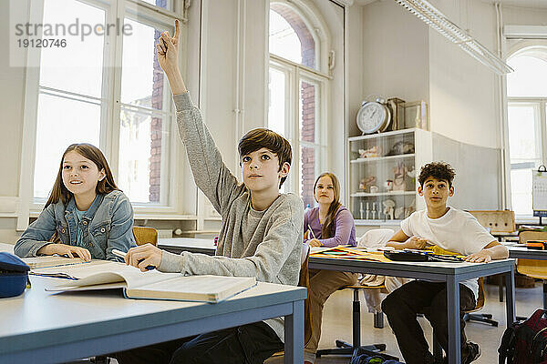Junge hebt die Hand  während er mit seiner Freundin am Schreibtisch im Klassenzimmer sitzt
