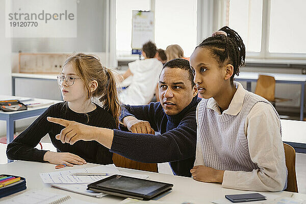 Männlicher Lehrer  der auf weibliche Schüler zeigt  die am Schreibtisch im Klassenzimmer sitzen