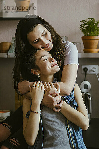 Lächelnde Frau  die ihre Freundin von hinten umarmt  während sie in der Küche zu Hause sitzt