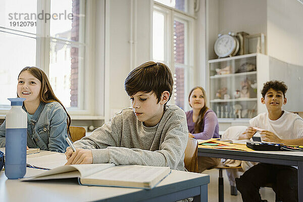 Schuljunge schreibt in ein Buch  während er mit Freunden im Klassenzimmer am Schreibtisch sitzt