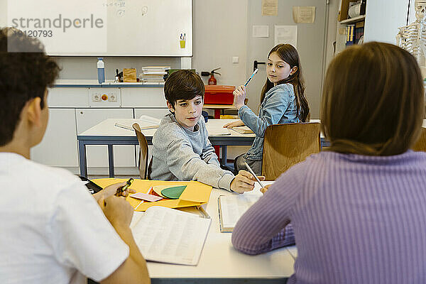 Junge nimmt Bleistift von einer Freundin  die am Schreibtisch im Klassenzimmer sitzt