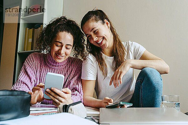 Lächelnde junge Frauen teilen sich ein Smartphone  während sie zu Hause mit Büchern sitzen