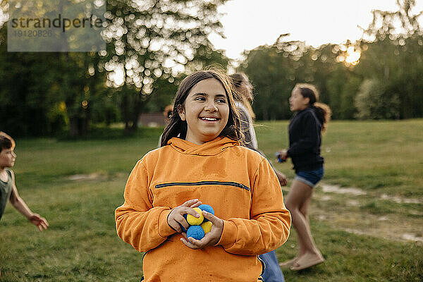 Lächelndes Mädchen  das blaue und gelbe Bälle hält  während es auf einem Spielplatz im Ferienlager steht