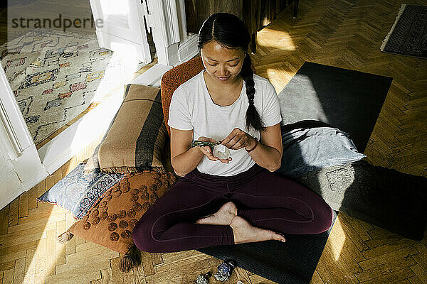 Hohe Winkel Ansicht der jungen Frau hält Kristall und Kraut beim Sitzen auf Übung Matte zu Hause
