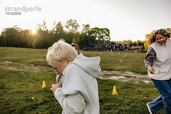 Seitenansicht eines blonden Jungen  der beim Spielen auf dem Spielplatz seinen Freunden ausweicht  die Wasser spritzen