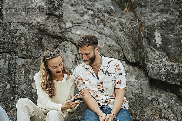 Lächelnde Frau  die ihr Smartphone mit einem männlichen Freund teilt  der vor einem Felsen sitzt