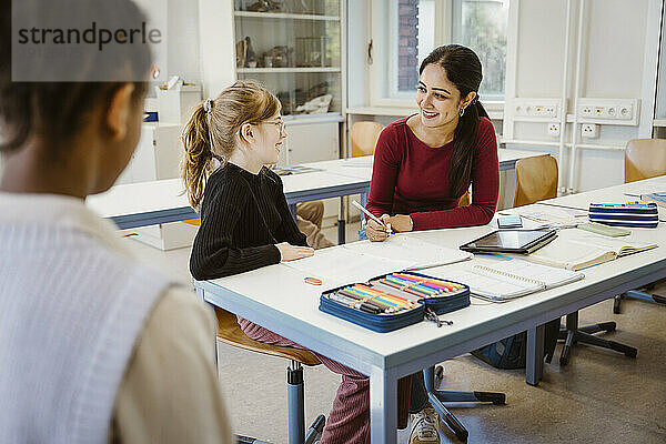 Lächelnde Lehrerin und Schülerin interagieren miteinander  während sie im Klassenzimmer sitzen