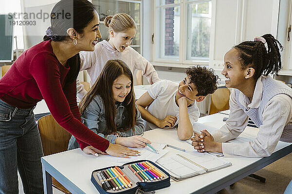 Glückliche Lehrerin lehnt am Schreibtisch mit Schülern im Klassenzimmer