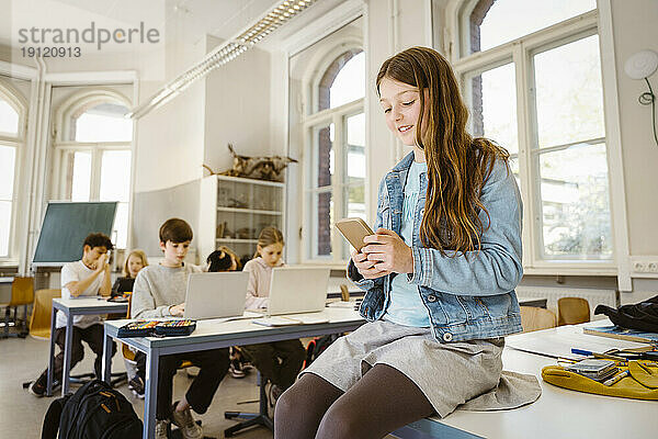 Lächelnde Studentin  die ein Smartphone benutzt  während sie am Schreibtisch im Klassenzimmer sitzt