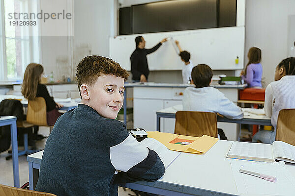 Porträt eines lächelnden Jungen  der über die Schulter schaut  während er auf dem Schreibtisch im Klassenzimmer sitzt