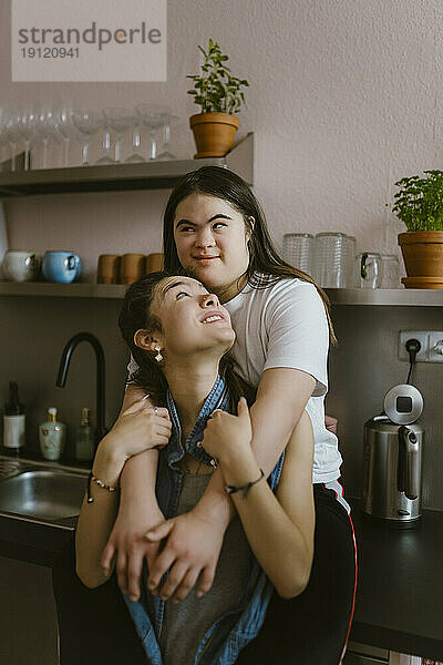 Junge Frau mit Down-Syndrom umarmt Schwester von hinten in der Küche zu Hause