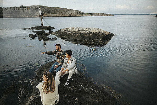 Hoher Blickwinkel auf einen Mann  der mit Freunden auf einem Felsen in der Nähe eines Sees sitzt und gestikuliert