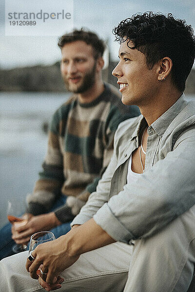 Junger Mann genießt Wein mit männlichem Freund am See sitzend