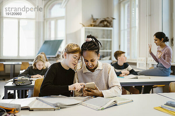 Neugierige Schulmädchen teilen sich ein Smartphone  während sie im Klassenzimmer am Schreibtisch sitzen