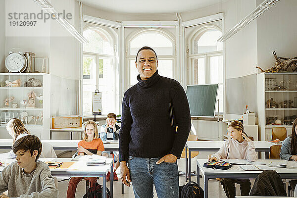 Porträt eines glücklichen männlichen Lehrers mit Hand in der Tasche  der inmitten von Schülern im Klassenzimmer steht