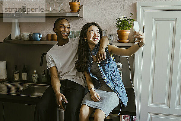 Glückliche junge Frau  die ein Selfie mit einem Freund macht  der auf der Küchentheke zu Hause sitzt