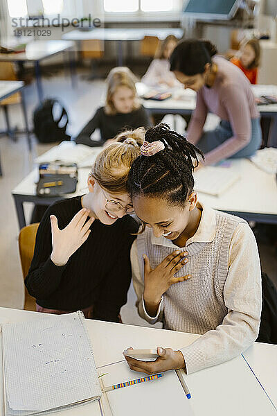 Hohe Winkel Ansicht der Schülerinnen gestikulieren  während mit Smartphone sitzen am Schreibtisch im Klassenzimmer