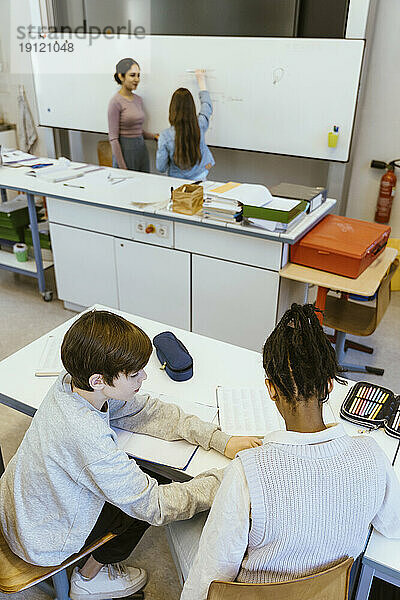 Hoher Blickwinkel eines Jungen  der einem männlichen Freund hilft  während er auf dem Schreibtisch im Klassenzimmer sitzt