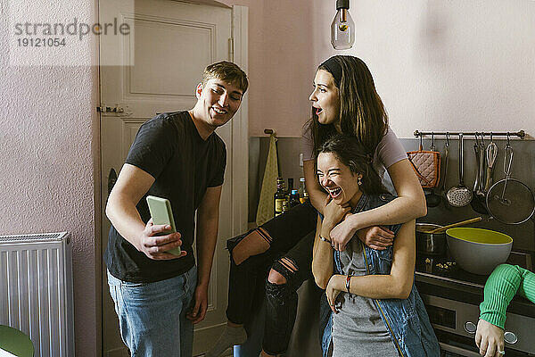 Lächelnder Mann teilt Smartphone mit fröhlichen Freundinnen in der Küche zu Hause