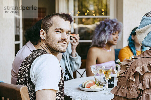Porträt eines lächelnden schwulen Mannes mit Freunden am Esstisch während einer Dinnerparty im Hinterhof