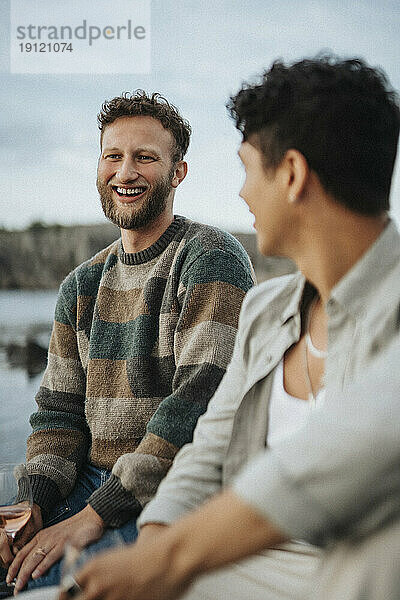 Glücklicher junger Mann genießt mit männlichen Freund in der Nähe von See sitzen