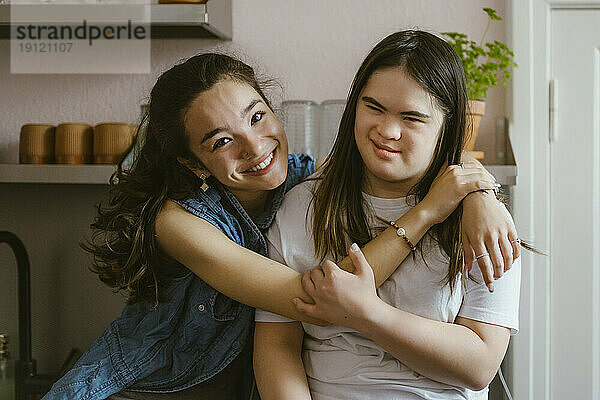 Porträt einer lächelnden jungen Frau  die ihre Schwester mit Down-Syndrom in der Küche zu Hause umarmt