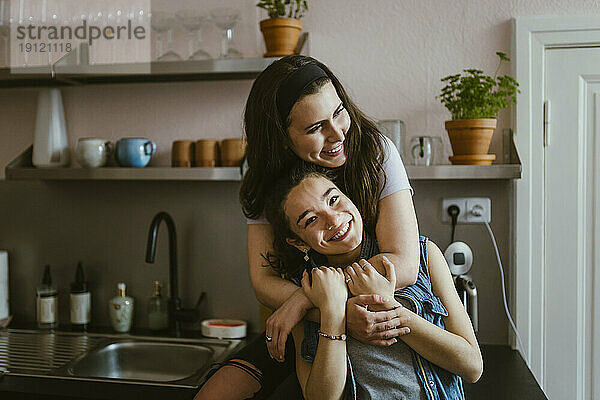 Porträt einer glücklichen Frau  die ihre Freundin von hinten umarmt  während sie zu Hause in der Küche sitzt