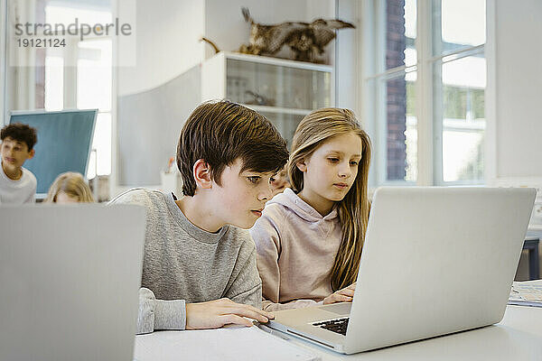 Konzentrierter Junge beobachtet Laptop mit Freundin am Schreibtisch im Klassenzimmer