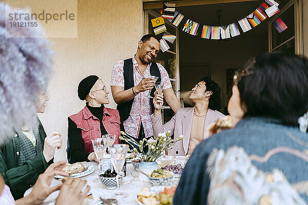 Lächelnder schwuler Mann genießt Getränke mit LGBTQ-Freunden während einer Dinnerparty im Hinterhof