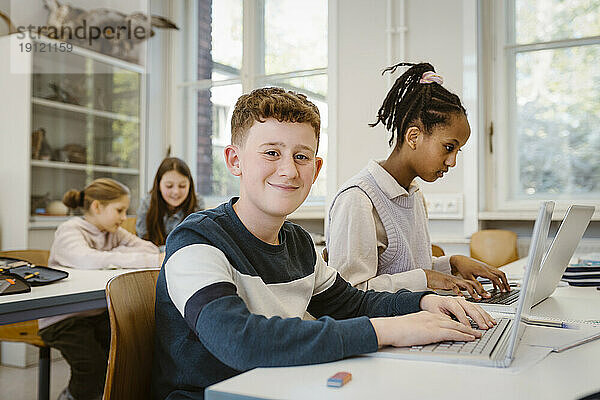 Porträt eines lächelnden Schülers mit Laptop bei einer Freundin im Klassenzimmer