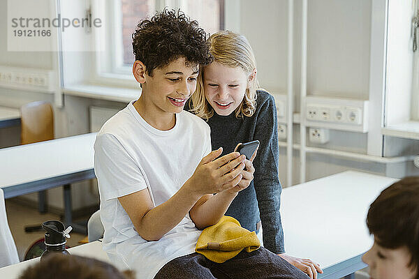 Neugieriger Junge und Mädchen teilen sich ein Smartphone  während sie am Schreibtisch im Klassenzimmer sitzen