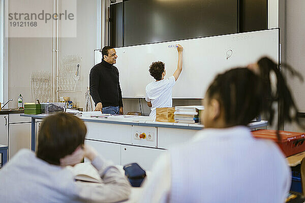 Lächelnde Lehrerin steht mit einem Schüler  der auf einer Tafel im Klassenzimmer schreibt