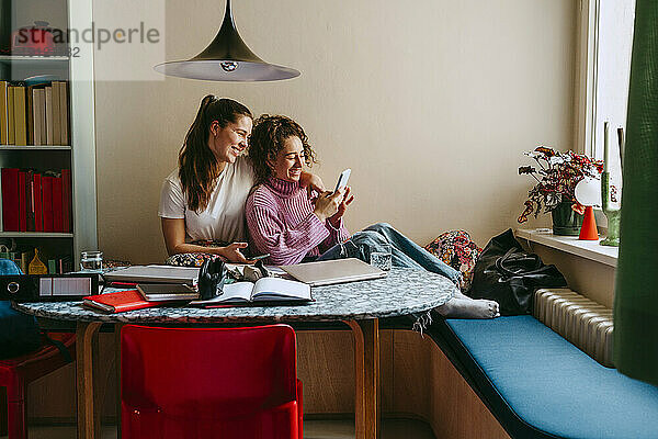 Glückliche junge Freundinnen teilen sich ein Smartphone  während sie zu Hause sitzen