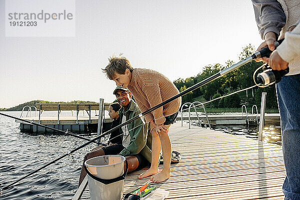 Neugieriger Junge  der in den See schaut  während ein Betreuer auf dem Steg im Sommerlager angelt