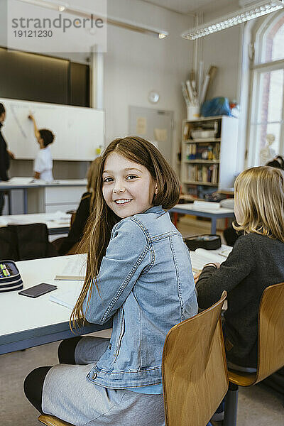 Seitenansicht Porträt eines lächelnden Mädchens  das eine Jeansjacke trägt und auf einem Stuhl im Klassenzimmer sitzt