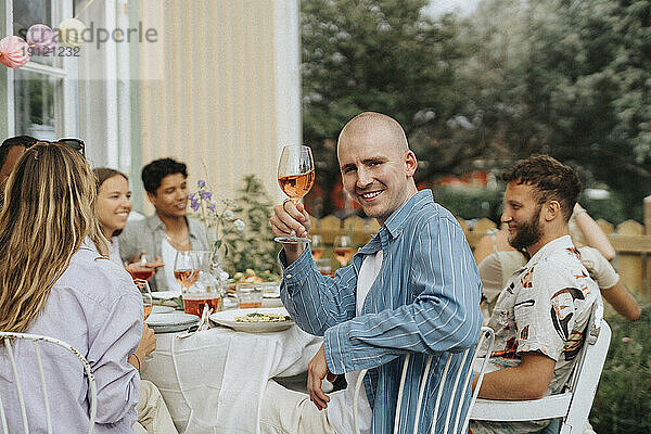 Seitenansicht Porträt eines lächelnden jungen Mannes  der ein Weinglas hält  während er mit Freunden auf einem Stuhl sitzt  während einer Dinnerparty im Café