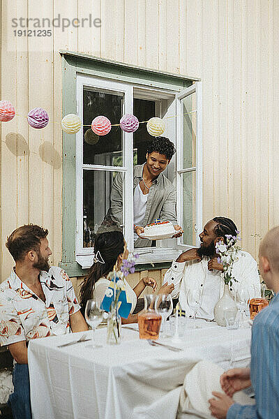 Lächelnder Mann  der Freunden am Tisch sitzend während einer Dinnerparty Kuchen schenkt