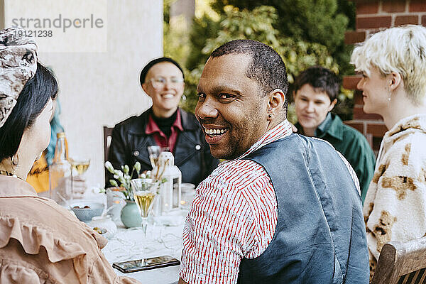 Porträt eines lächelnden schwulen Mannes  der Freunden bei einer Dinnerparty im Hinterhof über die Schulter schaut